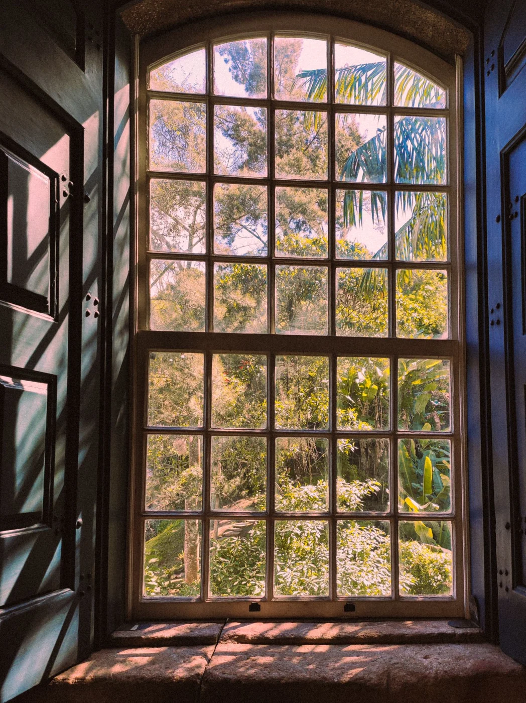 Sunlit garden view framed by a rustic arched window with interior wooden shutters.