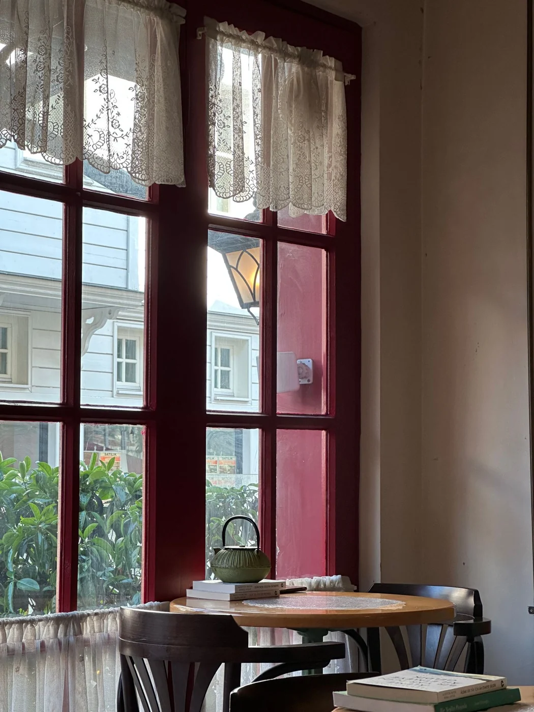 A serene view inside a cozy café with lace curtains and warm light streaming in through red-framed windows.