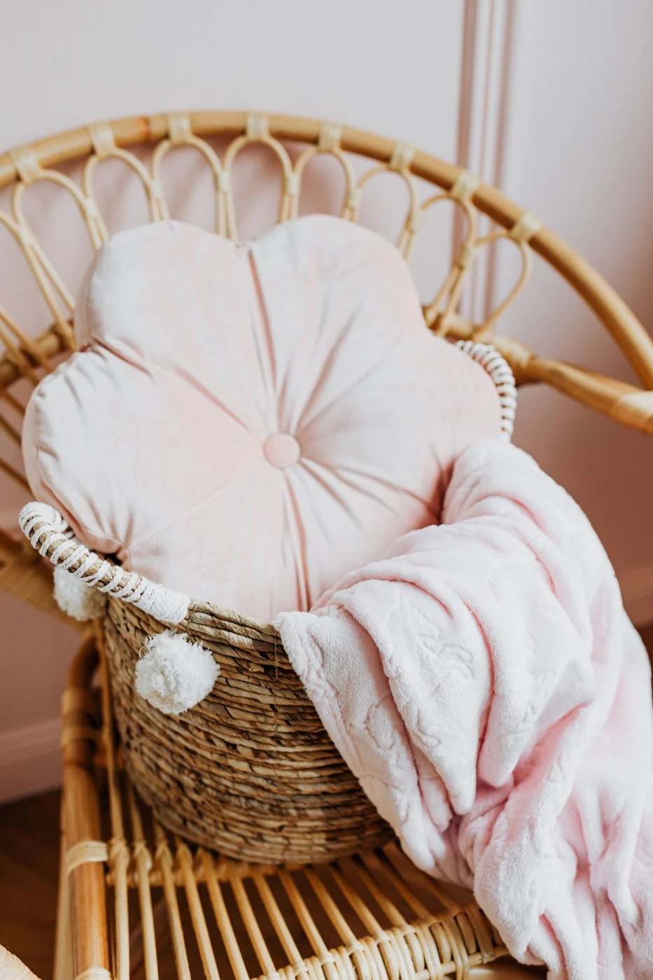 A stylish wicker chair holds a soft pink flower-shaped pillow and a plush blanket, adding a touch of cozy elegance.