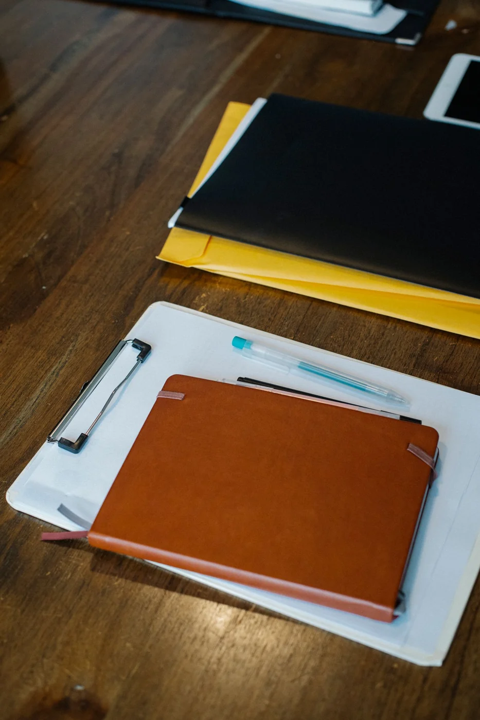 A notebook, clipboard and folders on a wooden desk.