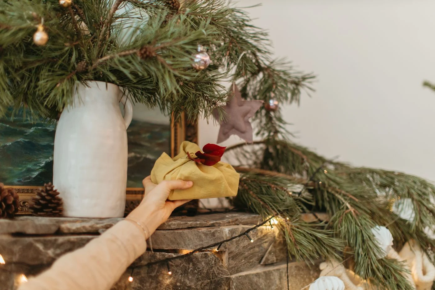 Hand holding a wrapped gift amidst Christmas decorations and greenery on a mantel.