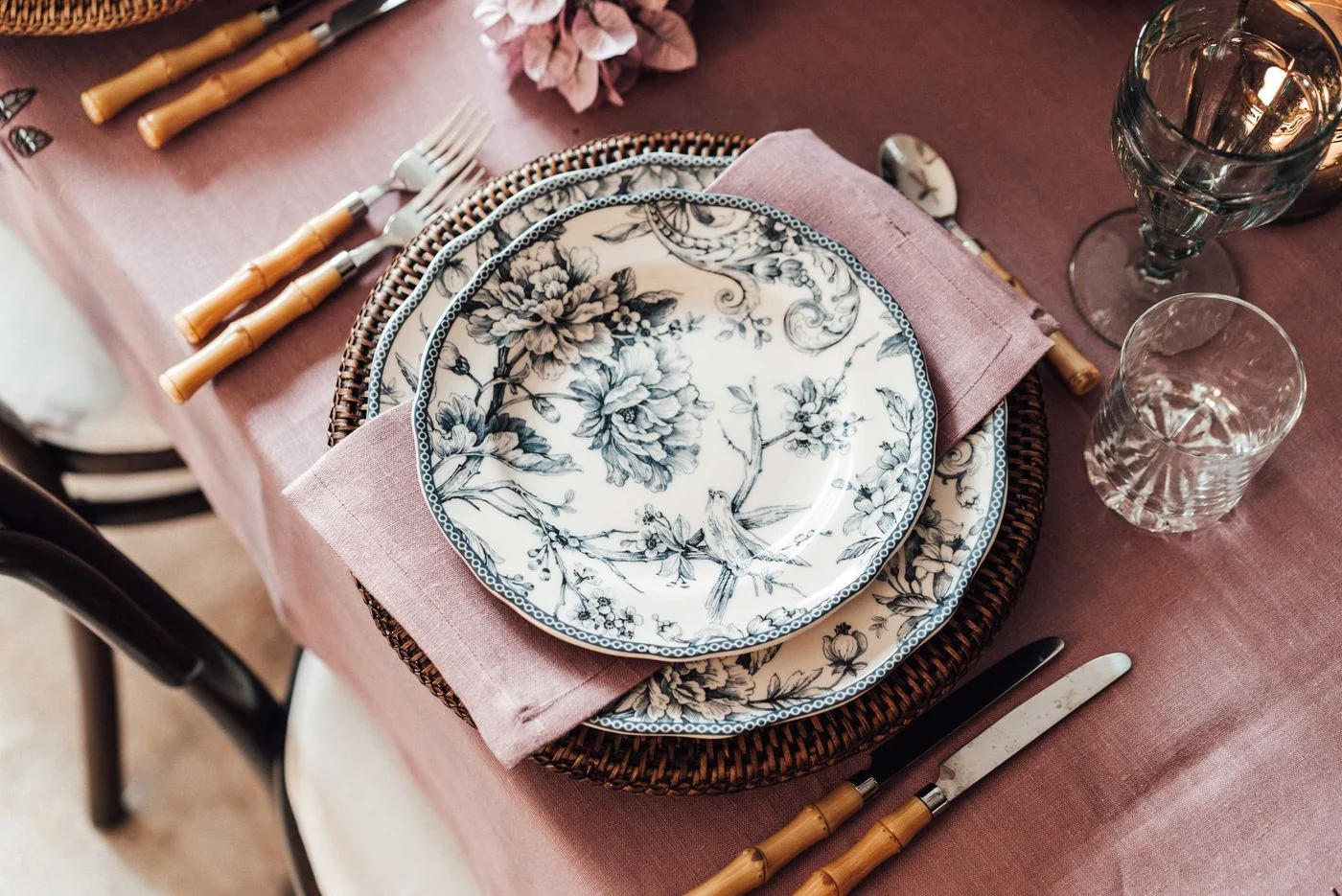 A sophisticated dinner table set with floral porcelain plates, bamboo cutlery, and pink linen napkin.