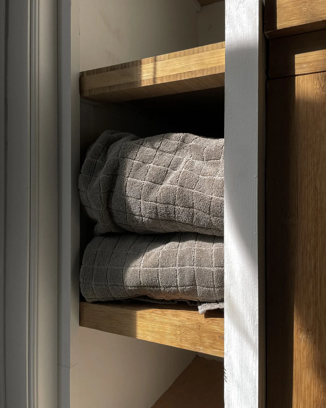 Soft, gray towels neatly folded in a sunlit wooden cabinet. Ideal for bathroom decor inspiration.