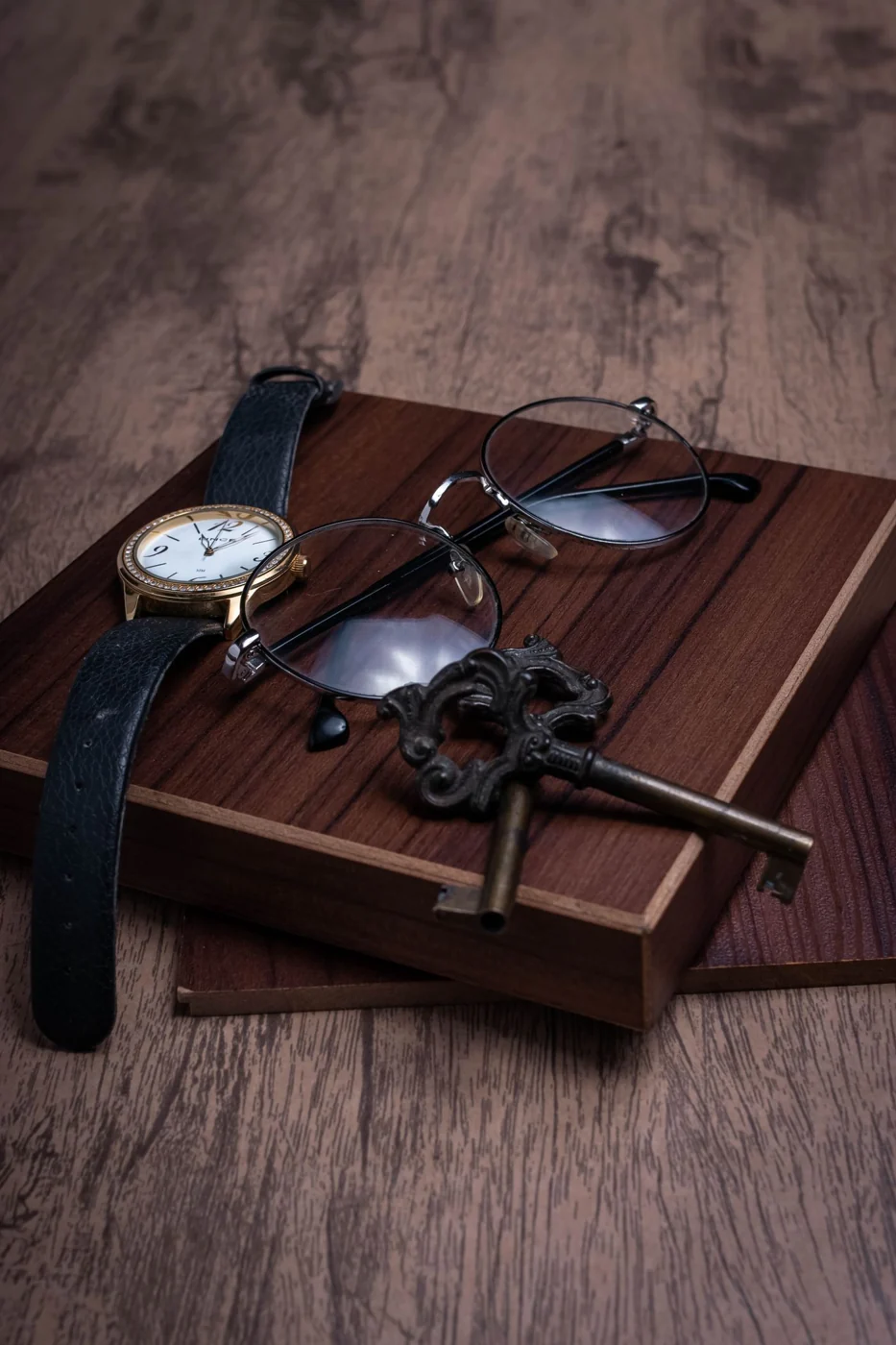 A vintage wristwatch, eyeglasses, and an antique key on a wooden tray, showcasing timeless elegance.