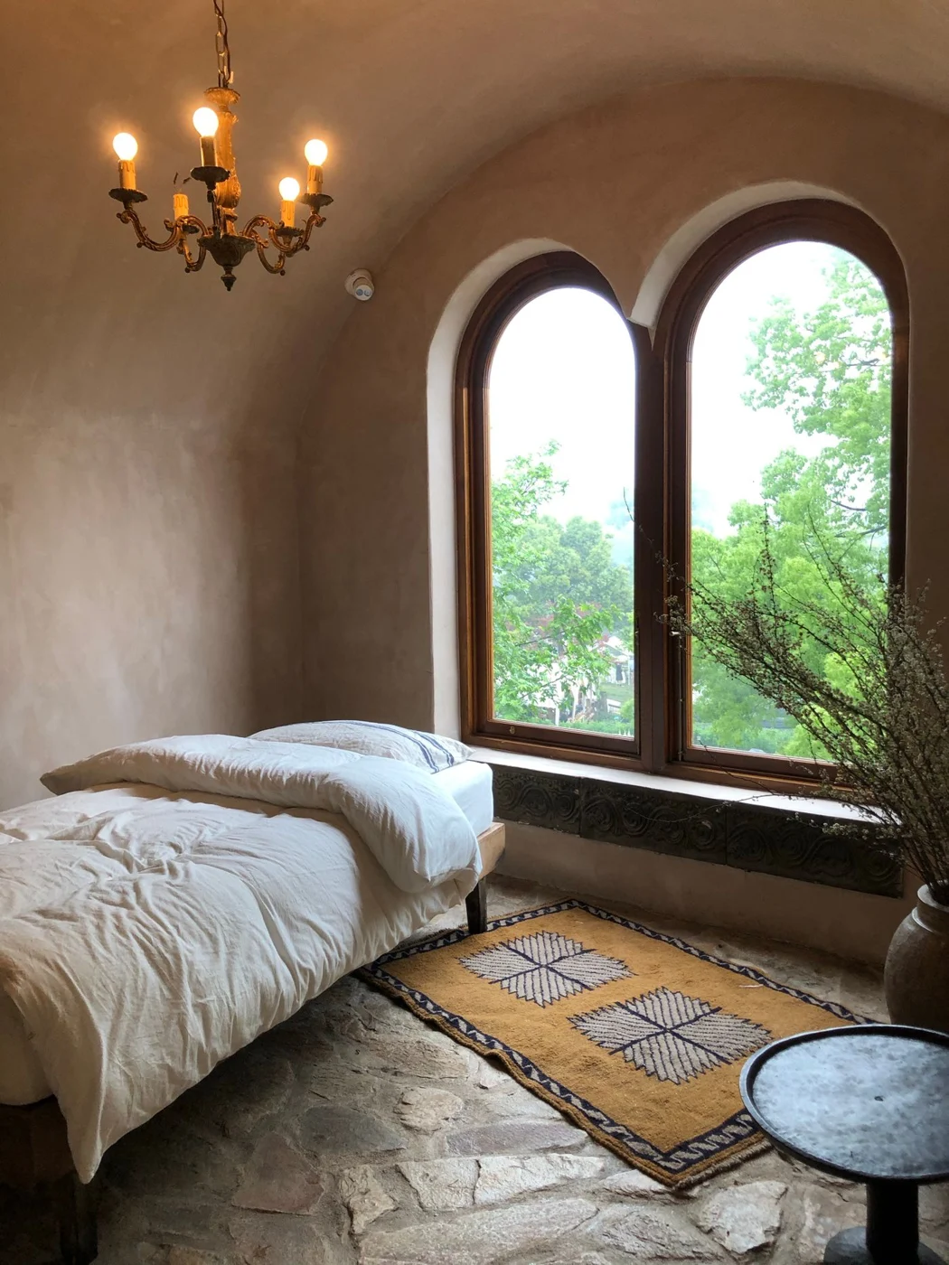 A rustic bedroom with irregular stone flooring, arched windows and pale bedding.